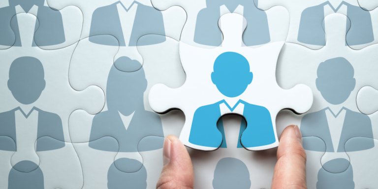 A hand placing the final puzzle piece into a jigsaw puzzle. The puzzle pieces feature silhouettes of business people, with the final piece showing a highlighted blue figure in a suit.